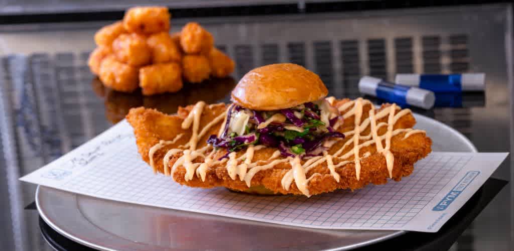 A plate features a crispy, oversized chicken cutlet topped with a small bun, coleslaw, and drizzle of sauce. In the background, a stack of golden tater tots adds a touch of comfort food appeal. The meal is presented on graph paper on a metal tray, giving a unique, playful vibe.