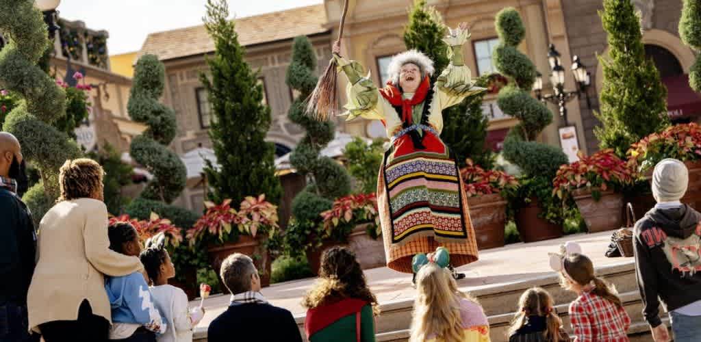 A performer in vibrant traditional attire entertains an engaged audience with lively gestures. The setting includes lush greenery and colorful decorations, creating a festive atmosphere. Adults and children sit and watch, captivated by the performance, in a beautifully landscaped outdoor venue.