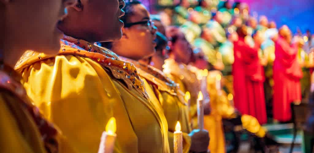A vibrant choir dressed in bright yellow robes holds candles while performing. The focus is on the singers, with blurred colorful lights and additional choir members in red robes in the background. The scene conveys a sense of harmony, unity, and celebration.