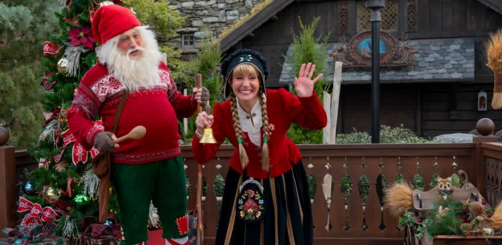 A cheerful holiday scene featuring a person dressed as Santa Claus next to a woman in traditional festive attire. They stand in front of a decorated Christmas tree, with a wooden house backdrop, creating a warm and welcoming festive atmosphere. Santa holds a bell and has a friendly expression, while the woman waves happily.