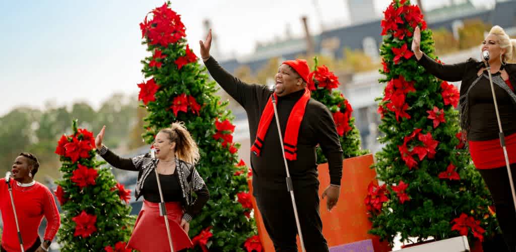 A group of performers sing joyfully outdoors, surrounded by festive decorations. They are dressed in red and black outfits, with vibrant poinsettia trees in the background. The atmosphere is lively and cheerful, capturing the essence of a holiday celebration.