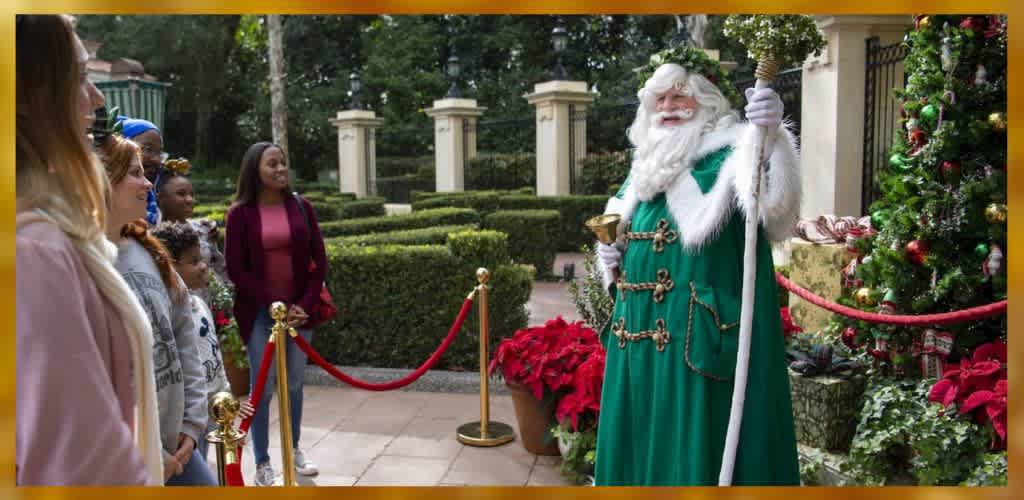 A festive scene shows a person dressed as Santa Claus in a green robe holding a bell and staff, standing next to a decorated Christmas tree surrounded by red poinsettias. They are engaging with a group of people including adults and children, behind a red rope barrier. The setting has manicured hedges and stone pillars, creating a holiday atmosphere.