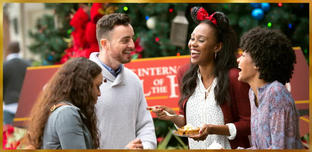 A group of four people stands in front of a festive holiday backdrop, featuring colorful decorations and a large sign. They are smiling and laughing, enjoying food together. The mood is joyful and celebratory, capturing the spirit of a fun and engaging social gathering. Holiday garb and bright colors enhance the festive atmosphere.