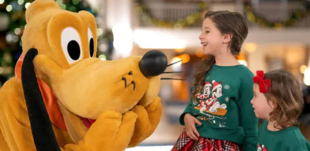A joyful moment at a holiday event features two young girls wearing festive sweaters with a dog character. The larger-than-life dog character is kneeling and playfully interacting with the smiling girls. The background is softly lit, with hints of holiday decor, creating a warm and cheerful atmosphere.