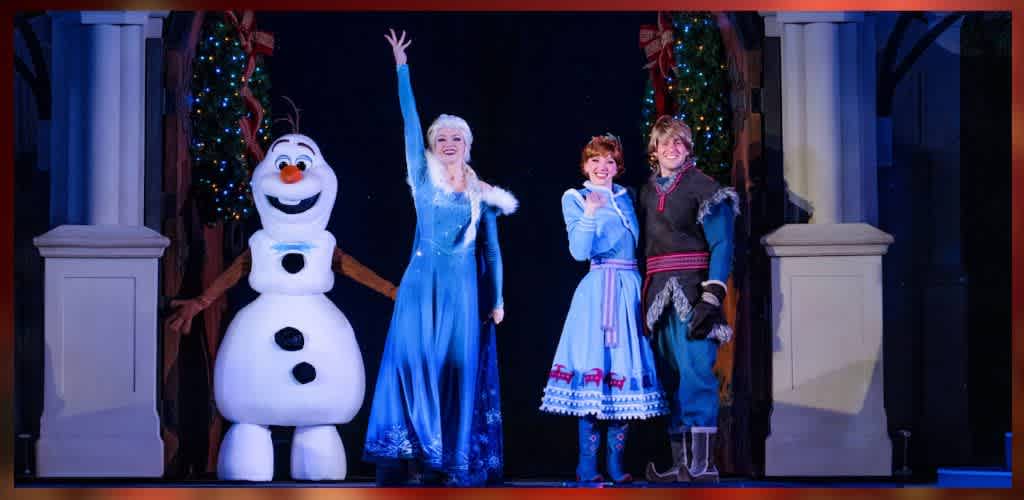 A joyful scene features a smiling snowman and three people on stage. The snowman is cheerful and round with black button-like details. Two women and a man stand beside it. One woman is in blue, raising her hand; the other woman is also in blue with braids, and the man is in a winter outfit. They are beneath a festive arch adorned with lights and greenery.