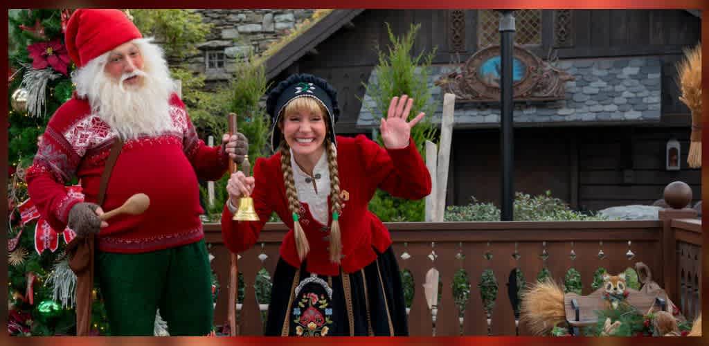 A festive scene with a person dressed as Santa Claus wearing a red hat and sweater, holding a wooden stick. Next to Santa, a smiling woman in a red and black traditional outfit waves and holds a bell. They are on a wooden platform with greenery in the background, giving a cheerful holiday vibe. There are also decorative elements like a red and green garland and small plush animals.