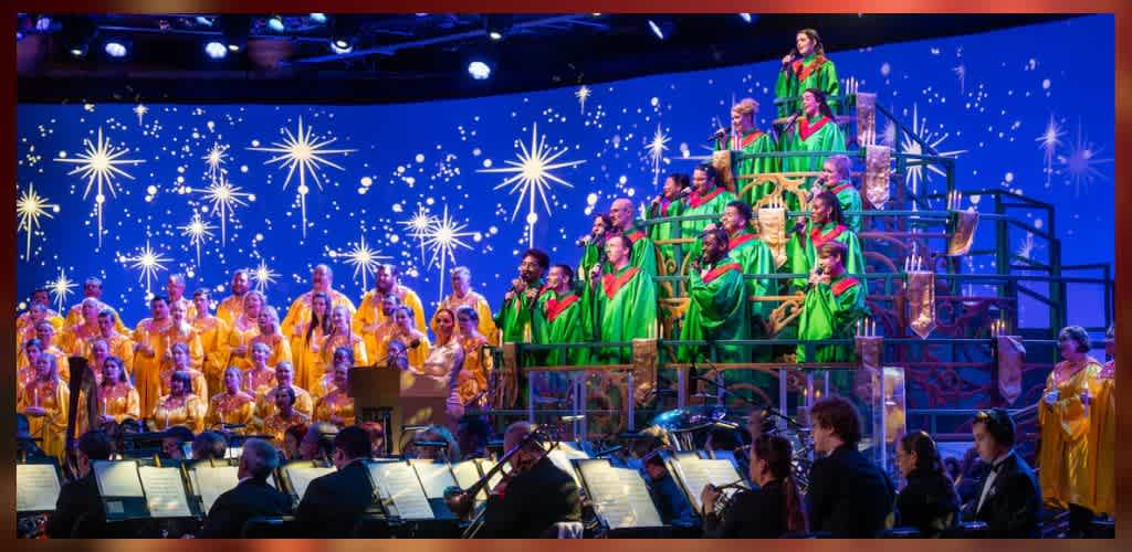 Choir performance featuring singers in green and yellow robes on tiered stands, set against a backdrop of twinkling stars on a blue screen. An orchestra plays in the foreground, enhancing the festive atmosphere. The scene is illuminated by stage lights, highlighting the colorful robes and vibrant setting.