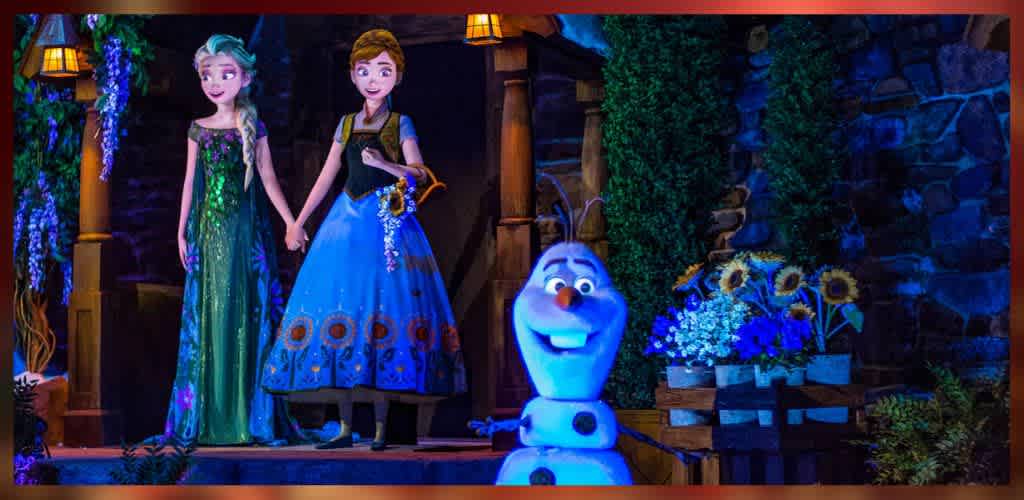 Two animated characters stand hand in hand on a stage. The character on the left wears a green gown, and the one on the right is in a blue dress adorned with floral patterns. In the foreground, a smiling snowman-like figure is visible. The background features a stone wall, wooden railings, and a variety of colorful flowers, creating a whimsical and enchanting atmosphere.