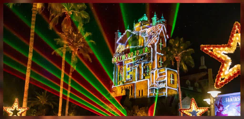 A nighttime scene featuring the Hollywood Tower Hotel illuminated with colorful projections and laser lights. Tall palm trees frame the building, enhancing the festive atmosphere. Bright stars made of lights decorate the foreground, contrasting against the dark night sky. The lively display creates a magical theme park environment ideal for nighttime entertainment.