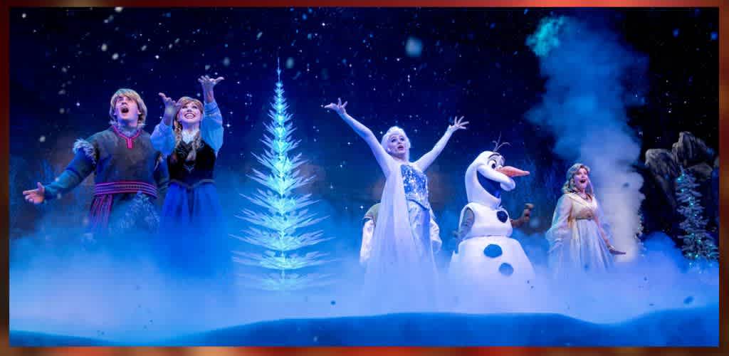 A group of animated characters celebrate joyfully in a snowy, magical setting. A glowing tree stands in the center, with sparkling snowflakes falling around them. The scene features bright costumes and expressions of happiness, creating a vibrant winter wonderland atmosphere.