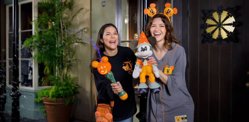Two women stand smiling at a theme park with festive, themed merchandise. Both wear themed ears and hold playful items, including a bubble wand and a stuffed toy. One is dressed in a dark sweater while the other wears a gray outfit. The background features greenery and a decorative door, adding to the joyful atmosphere. Bubbles float around, enhancing the celebratory feel.