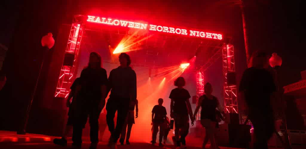 People walk toward a stage with red lighting and a sign that says Halloween Horror Nights at night.