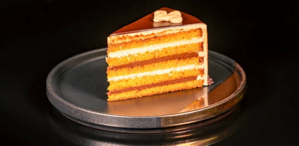 A slice of layered caramel cake with white frosting and a decorative topping on a glass plate against a black background