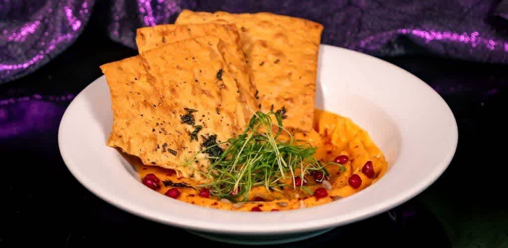 Grilled flatbread served with hummus garnished with microgreens and pomegranate seeds on a white plate with a dark background.