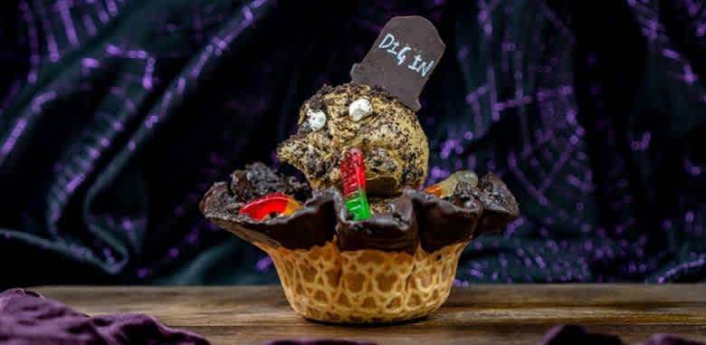 A Halloween themed dessert with a waffle bowl filled with chocolate and candy, featuring a cookie decorated as a spooky creature with a dig in sign and colorful candy.