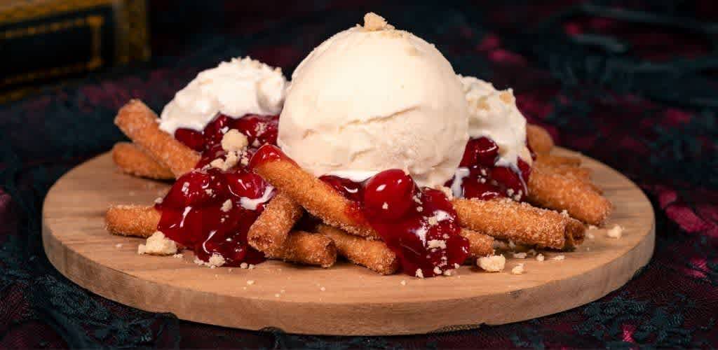 A dessert with vanilla ice cream, cherry topping, crispy fried sticks, and crumbles served on a round wooden platter.