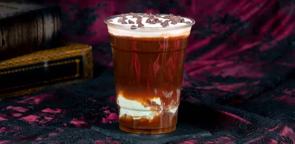 A layered chocolate and vanilla dessert in a clear glass topped with chocolate shavings on a dark fabric background.