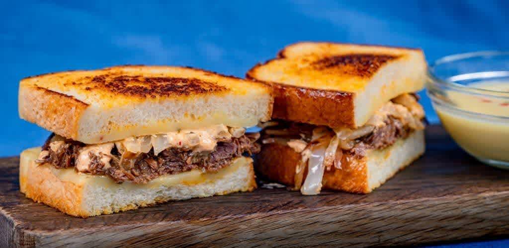 Two grilled sandwiches with shredded beef and coleslaw on toasted bread, served on a wooden surface with a side of dipping sauce.