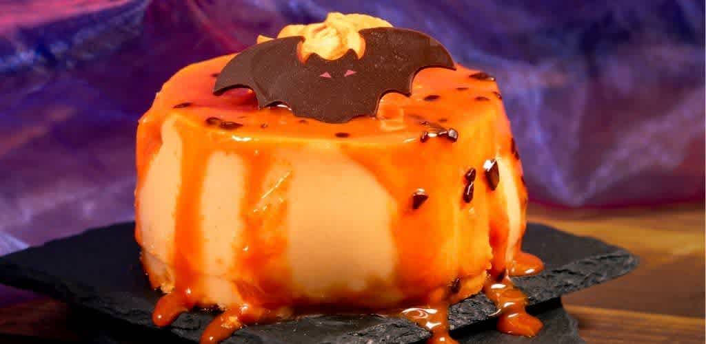 A Halloween-themed caramel cheesecake topped with a bat decoration, drizzled with caramel sauce on a black slate platter.