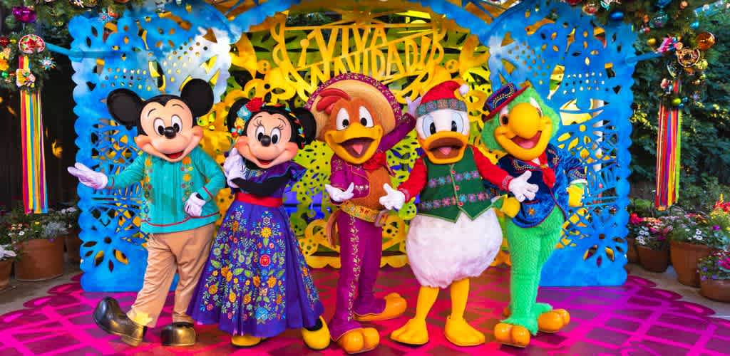 Five costumed Disney characters including Mickey Mouse, Minnie Mouse, Daisy Duck, Donald Duck, and Goofy standing together in front of colorful holiday decorations outside.