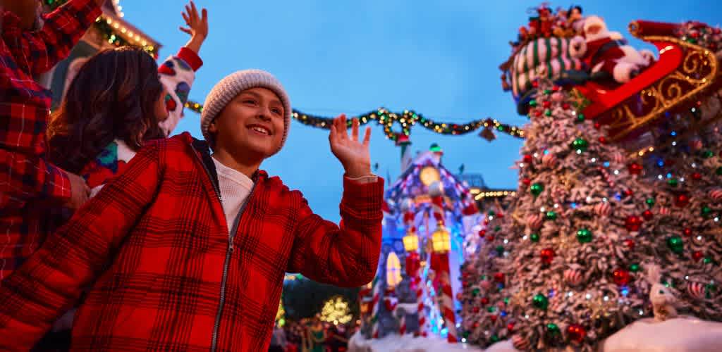 Children smiling and waving at a festive outdoor Christmas light display with decorated trees and colorful lights at dusk