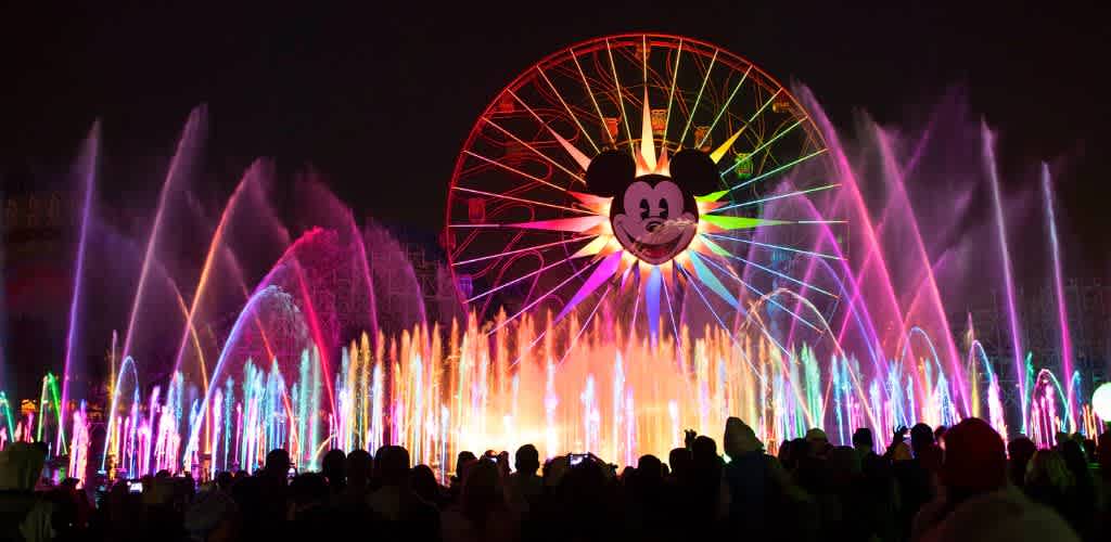 Famous Mickey Mouse face displayed on a large Ferris wheel with colorful fountain lights and water sprays at night.