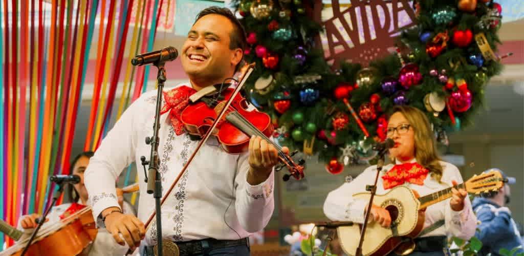 Musicians performing with a violin and guitar in front of a decorated Christmas tree and colorful ribbons at a festive event