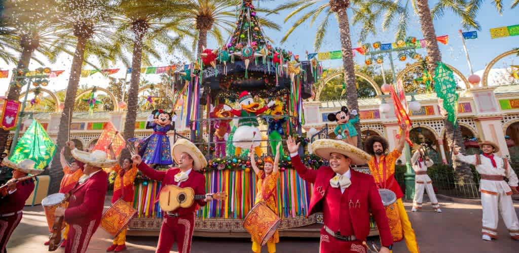 Colorful parade with musicians, dancers, and Disney characters celebrating around a decorated float with palm trees and festive banners under a sunny sky