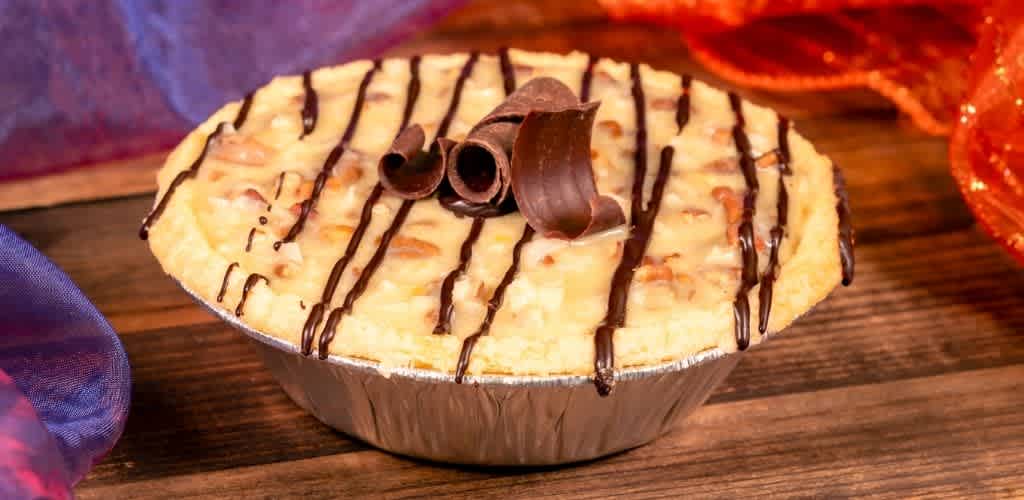 A baked cheesecake in a foil cup topped with chocolate shavings and drizzled with chocolate sauce on a wooden surface.