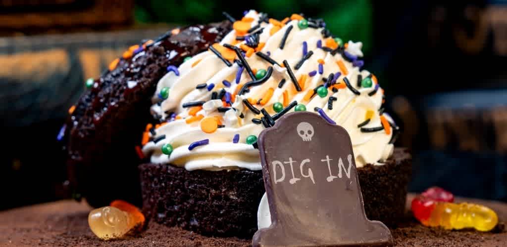 A chocolate cupcake topped with white frosting colorful sprinkles and candies inside a bakery setting with a Halloween themed sign reading Dig In