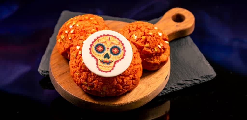 Three sugar cookies with orange icing and sprinkles on a wooden cutting board, one decorated with a colorful sugar skull design for Dia de los Muertos celebration.