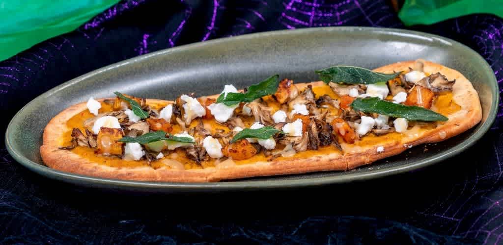 A flatbread pizza topped with roasted vegetables, fresh green herbs, and crumbled cheese served on a green oval plate.