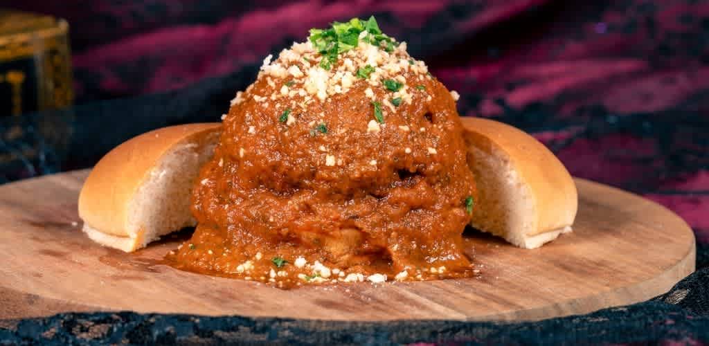 A serving of chili on a wooden board with bread rolls, topped with cheese and green herbs, against a dark, colorful background.