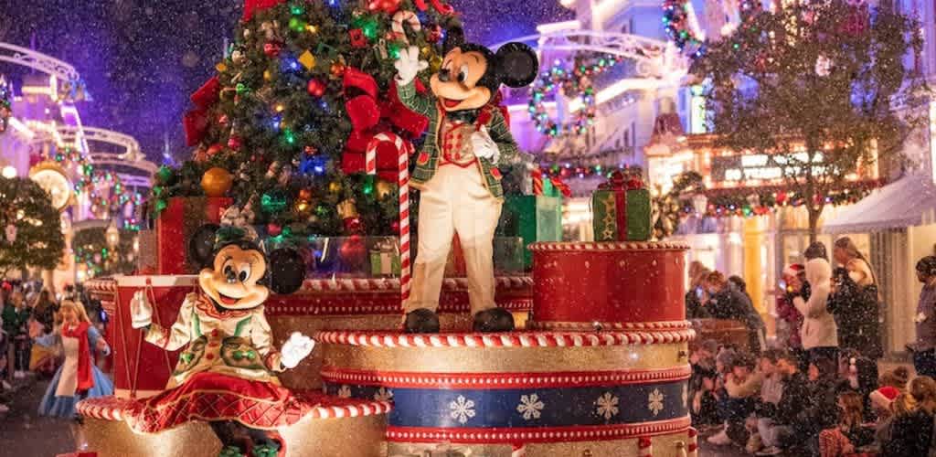 Festive holiday scene featuring Mickey Mouse and Minnie Mouse dressed in Christmas attire on a decorated parade float with a Christmas tree and surrounded by holiday lights and spectators