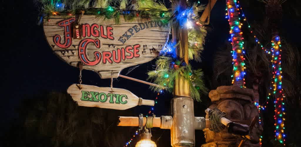 A wooden sign advertising Jingle Cruise with colorful holiday lights, greenery, and tropical decorations against a dark night sky.