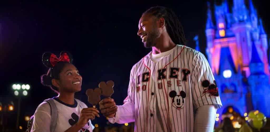 A man and a girl with Mickey Mouse themed accessories sharing Disney themed Mickey Mouse shaped ice creams at night with a castle in the background.