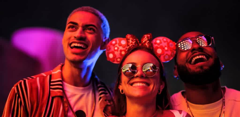Three people smiling happily at a colorful party, wearing fun accessories and glowing in vibrant lighting.