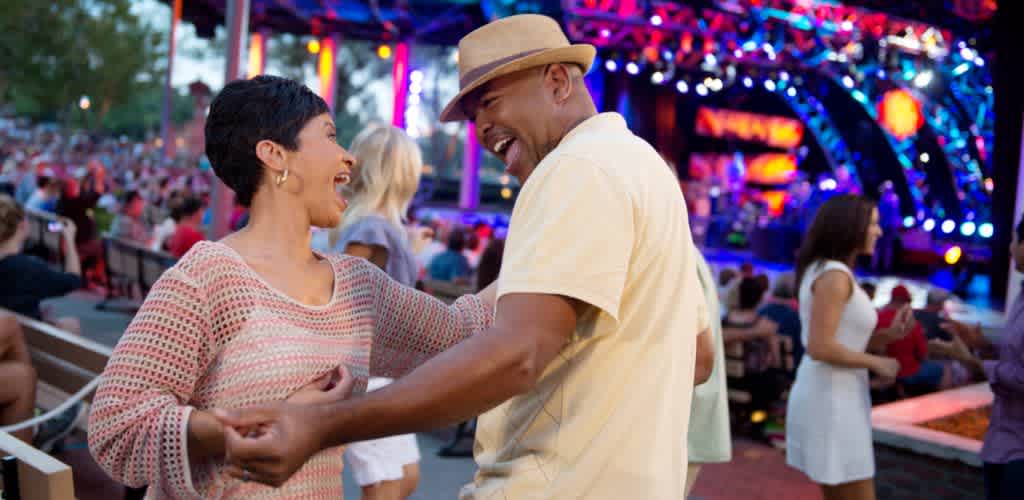 Couple dancing outdoors at a lively music festival with vibrant stage lights and a crowd in the background