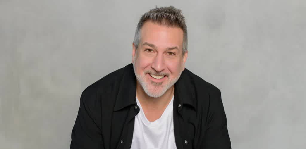 A smiling middle-aged man with gray hair and beard wearing a black jacket and white shirt sitting against a neutral background.