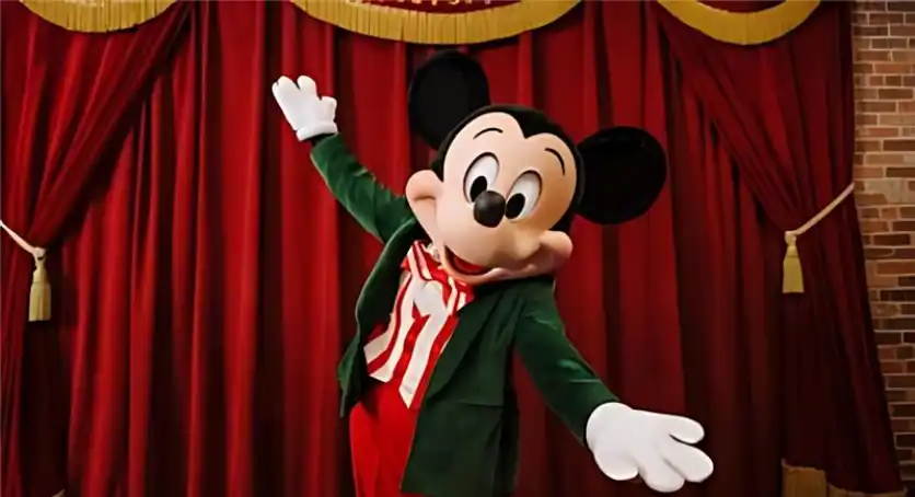 Mickey Mouse wearing a green holiday jacket and red-striped shirt, posing cheerfully in front of a red velvet stage curtain at Disneyland.