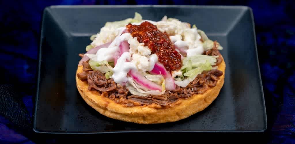 A tostada topped with shredded beef, lettuce, onions, sour cream, cheese, and salsa served on a black square plate