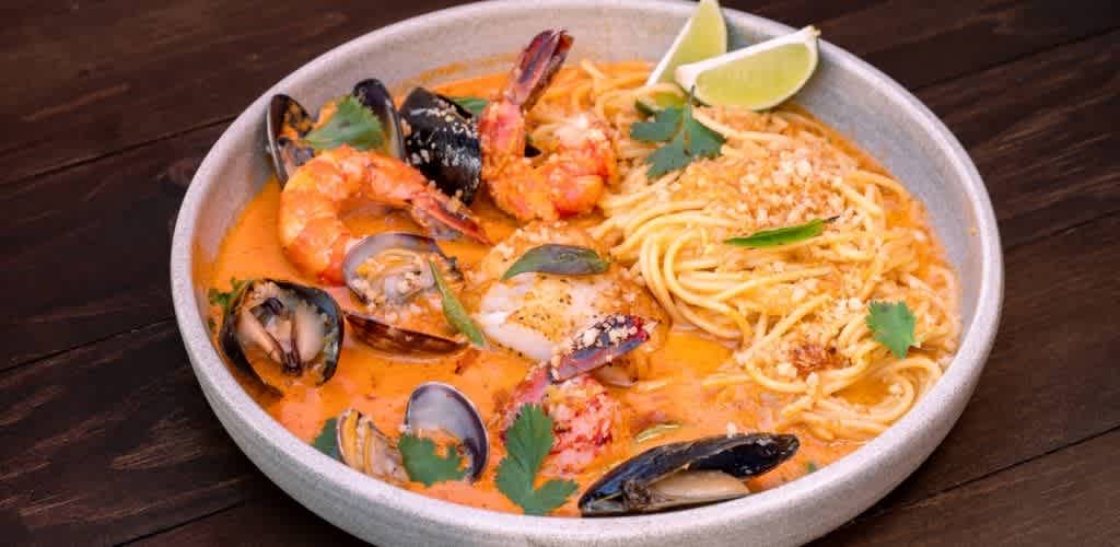 A bowl of seafood pasta with shrimp, mussels, and spaghetti in a creamy orange sauce garnished with lime wedges, cilantro, and grated cheese.