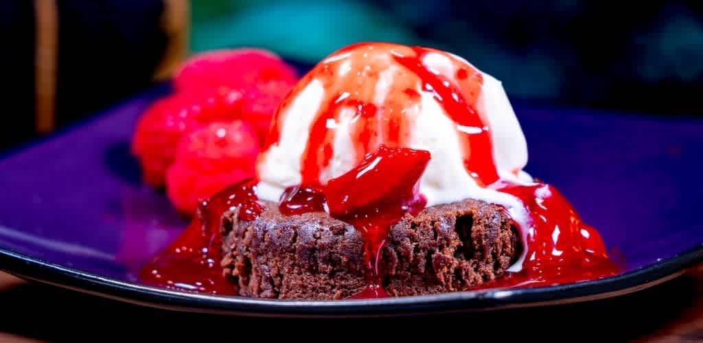 Chocolate brownie topped with vanilla ice cream and strawberry syrup with fresh strawberries on the side on a dark plate