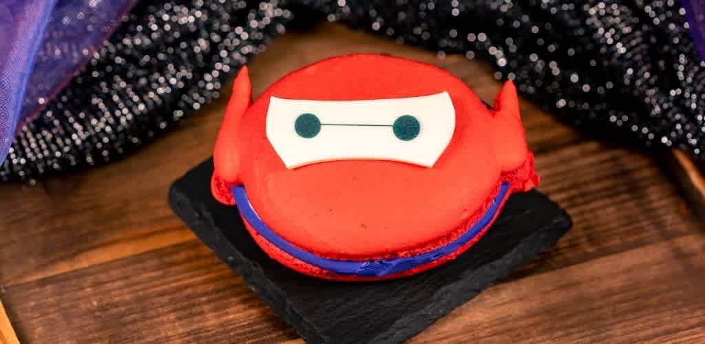 A round red cake decorated with white and black fondant to resemble a cartoon superhero character with a smiling face and a black base.