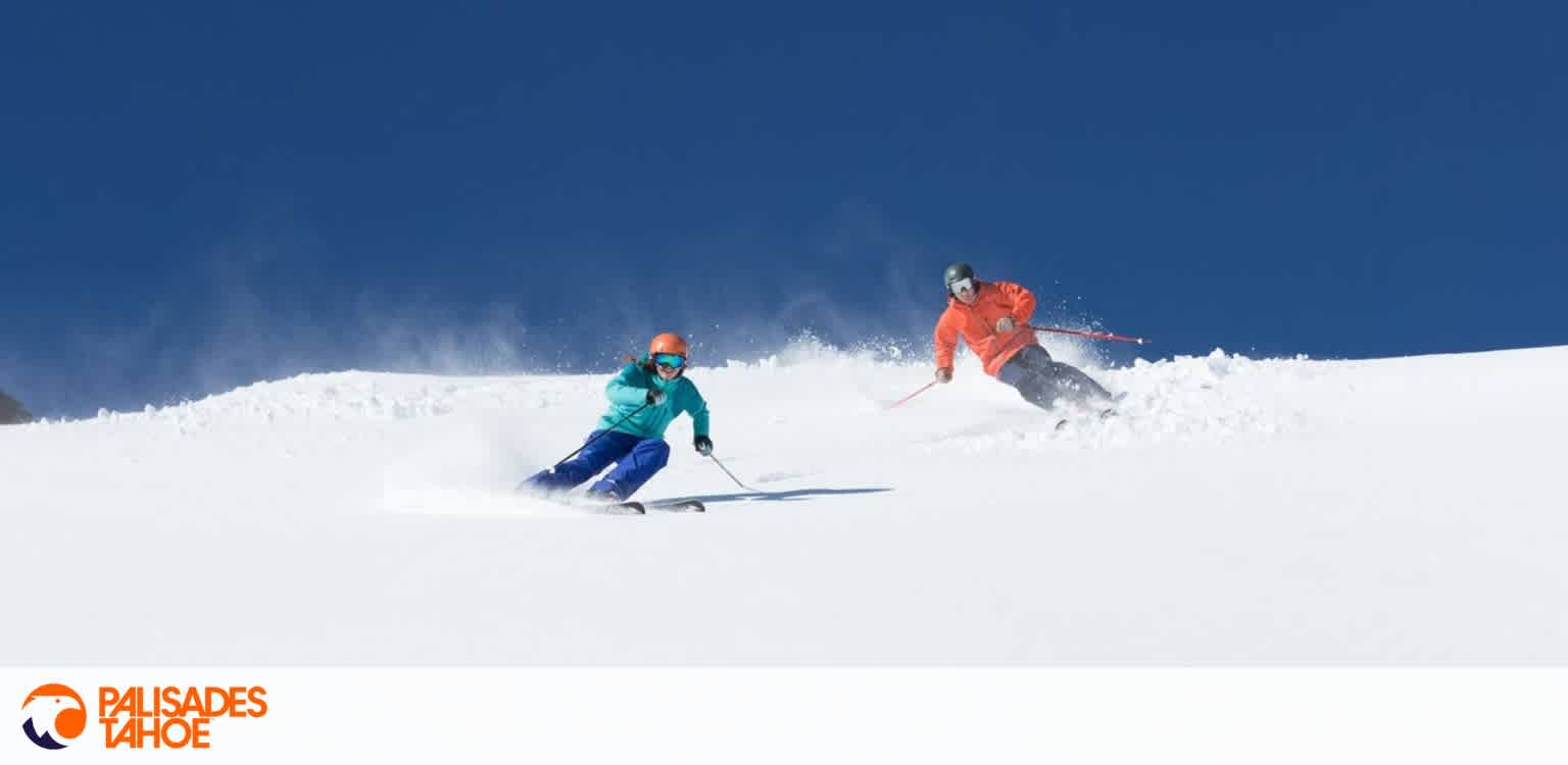 Two skiers in bright outdoor gear skiing down a snowy slope under a clear blue sky.