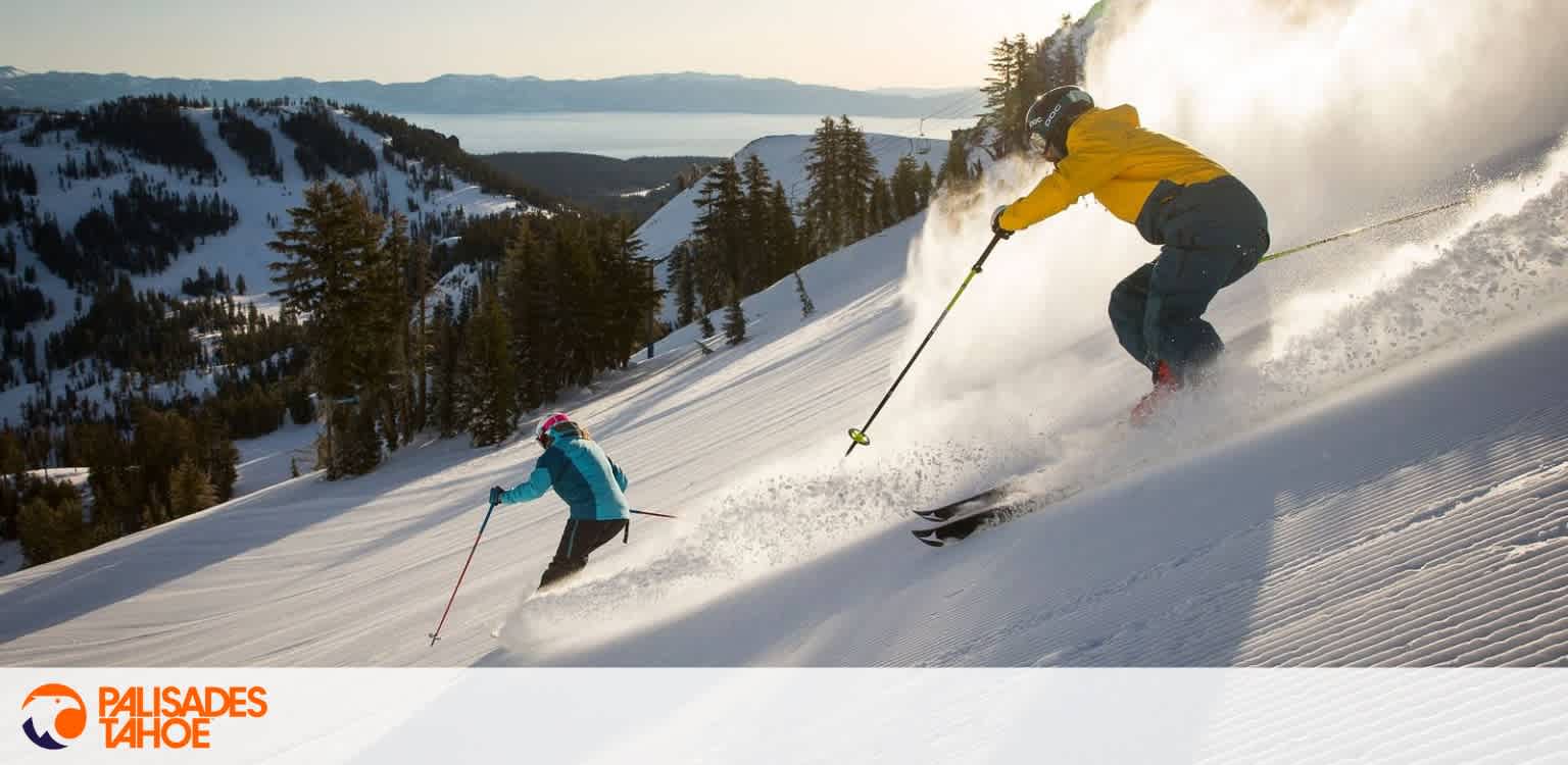 Two skiers in colorful gear descend a snowy mountain slope with pine trees and a distant lake in the background