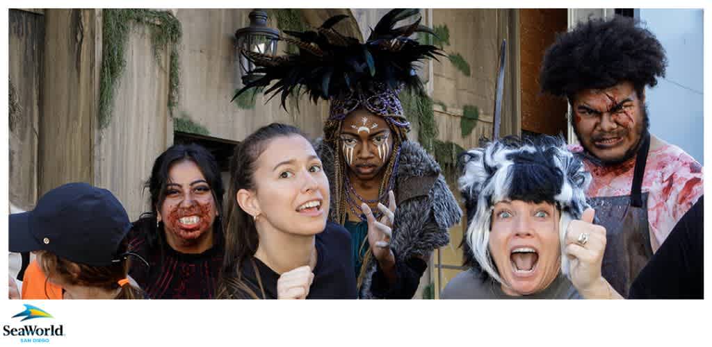A group of people in costumes and makeup with varied expressions, including surprise and intimidation, posing in front of a concrete wall with greenery.