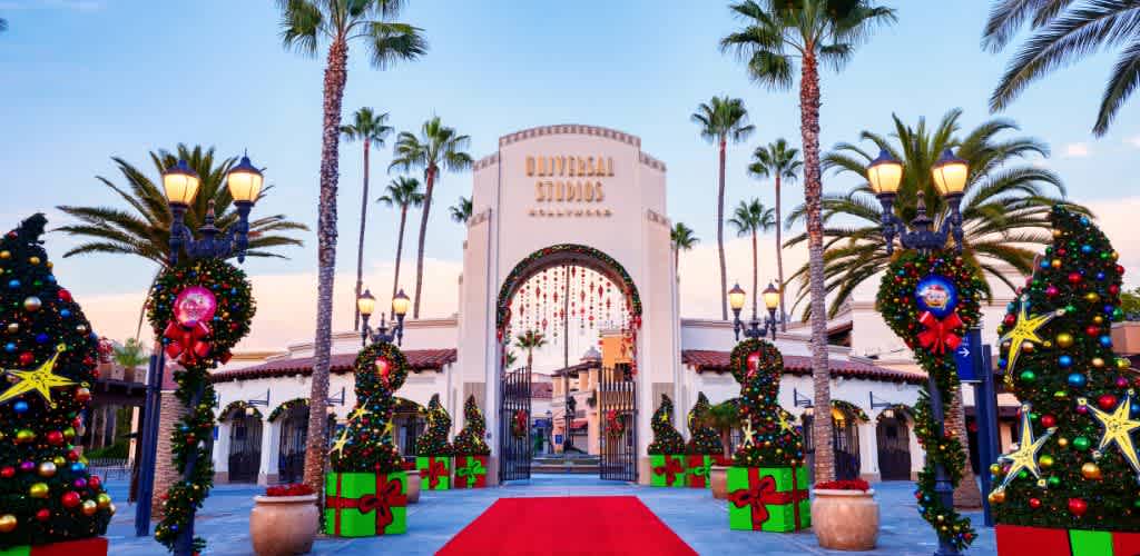 Entrance to a theme park bordered by tall palm trees and classic street lamps. A vibrant red carpet leads to an archway that reads Universal Studios Hollywood. The sky is clear and blue, adding to the lively atmosphere.