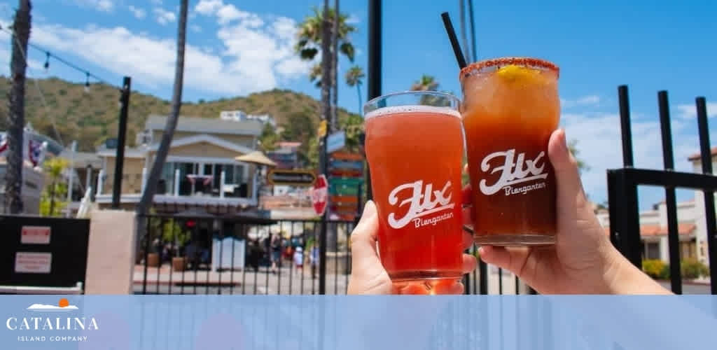 Two hands holding drinks in front of a sunny outdoor scene with palm trees and a town backdrop.
