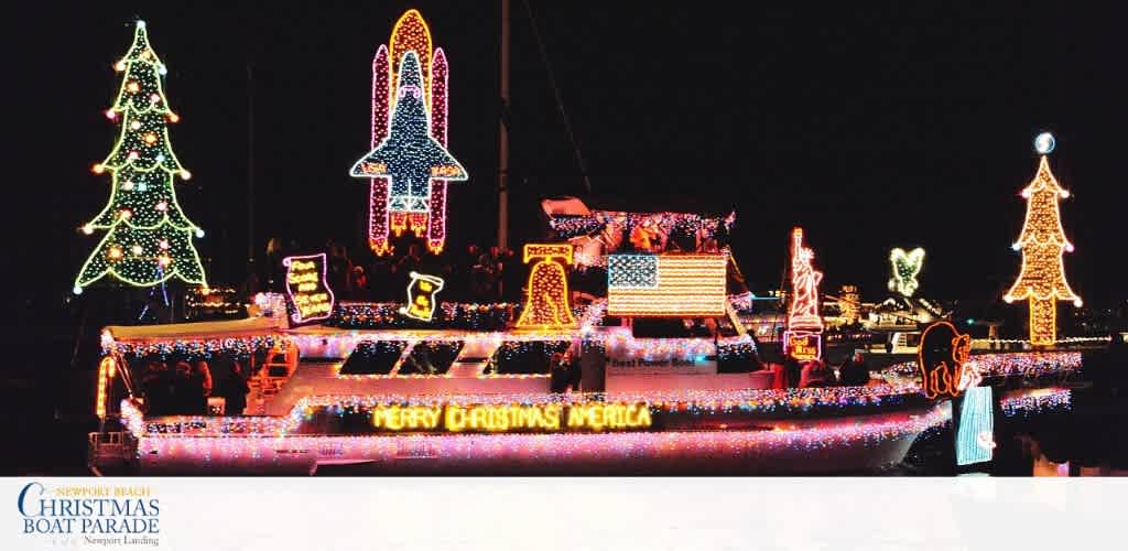 A boat decorated with colorful Christmas lights, trees, ornaments, and a sign that says Merry Christmas America during a nighttime parade.